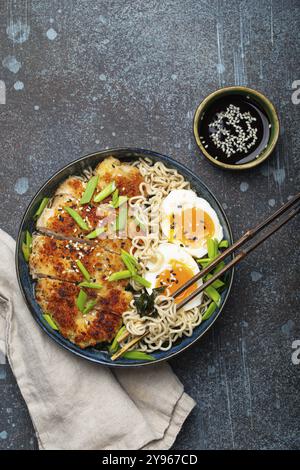 Zuppa di ramen di pasta asiatica con filetto di pollo fritto al panko e uova sode in un recipiente di ceramica con bastoncini di tritare e salsa di soia su backgroun rustico di pietra Foto Stock