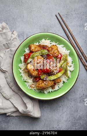 Pollo asiatico agrodolce e appiccicoso con verdure, friggere e riso in una ciotola di ceramica con bacchette, vista dall'alto su fondo in pietra rustica grigia, mestiere Foto Stock