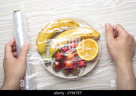 Mani maschili che avvolgono vari tipi di frutta e frutti di bosco su un piatto bianco in pellicola trasparente in plastica vista dall'alto su un tavolo rustico in legno bianco, cibo p Foto Stock