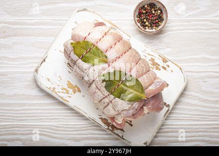 Carne di maiale a fette arrotolata, con foglie di alloro e condimenti su un tagliere bianco su sfondo rustico in legno bianco. Involtino di maiale pronto per essere consumato Foto Stock