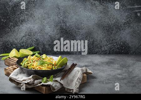 Tagliatelle vegetariane asiatiche con verdure e lime in una ciotola nera in ceramica rustica, bacchette di legno sul tagliere vista angolare su fondo di pietra. COO Foto Stock