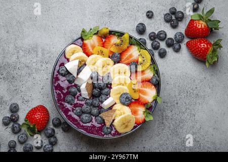 Frullato estivo di acai salutare con semi di chia, banana fresca, fragole, mirtilli, cocco, vista dall'alto kiwi su sfondo rustico in cemento con cucchiaio Foto Stock