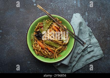Piatto asiatico mescolare gli spaghetti di udon fritti con pollo, verdure e funghi in una ciotola di ceramica verde con bacchette di legno sul retro rustico di cemento blu scuro Foto Stock