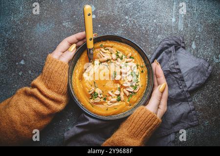 Mani femminili in maglione a maglia giallo che tiene una ciotola con zuppa di crema di zucca su fondo di pietra scura con cucchiaio decorato con arachidi, vista dall'alto. Au Foto Stock