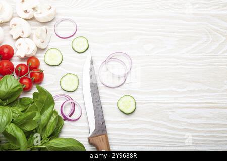 Cucina di base: Ingredienti freschi con coltello da cucina, basilico, pomodori ciliegini, funghi, cetriolo tagliato e cipolla su sfondo di legno bianco a. Foto Stock