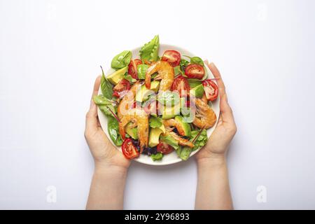 Mani femminili che tengono sana insalata con gamberetti grigliati, avocado, pomodori ciliegini e foglie verdi su piatto bianco isolato su fondo bianco vie Foto Stock