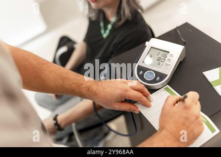 Primo piano delle mani di un medico di sesso maschile che scrive le misure della pressione sanguigna femminile da un dispositivo digitale in farmacia Foto Stock