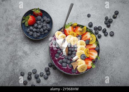 Frullato estivo di acai salutare con semi di chia, banana fresca, fragole, mirtilli, cocco, vista dall'alto kiwi su sfondo rustico in cemento con cucchiaio Foto Stock