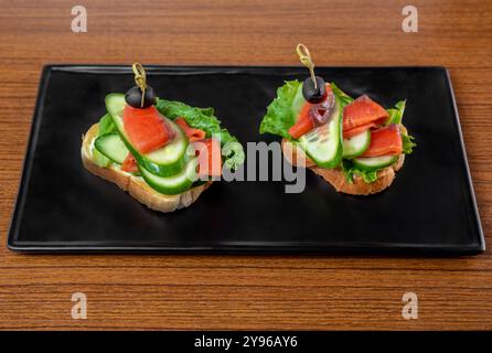 panini con una fetta di tenero salmone affumicato e verdure su un piatto nero su un tavolo di legno. Foto Stock