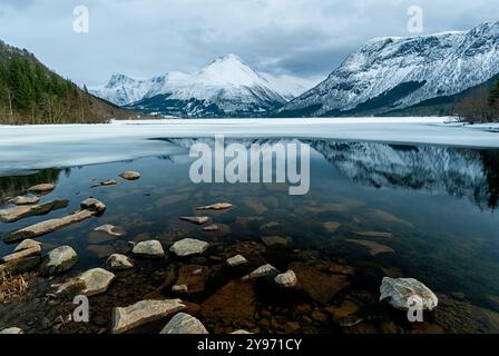 Una vista tranquilla delle acque tranquille che mostra il riflesso delle maestose montagne sotto un cielo nuvoloso, circondato da ghiaccio e alberi. Foto Stock