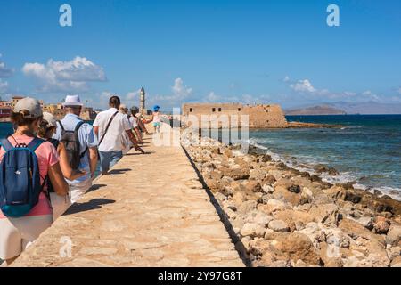 Turismo Europa, vista posteriore di un gruppo di turisti a Chania, Creta, camminando in un unico file sul vecchio porto veneziano alle mura del mare verso un faro. Foto Stock