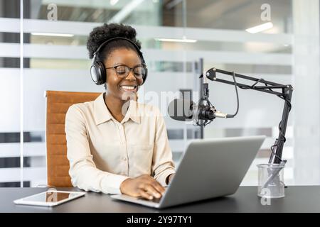 Donna sorridente che registra podcast in un ufficio moderno con laptop, microfono e cuffie. Impegnata nella creazione di contenuti digitali, discutendo argomenti con il pubblico. Atmosfera calma, professionale e concentrata. Foto Stock