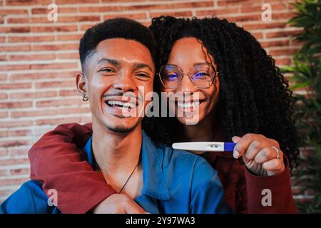 Coppia afroamericana celebra un test di gravidanza positivo a casa. Abbracciano e sorridono, condividendo l'eccitante notizia di aspettarsi un bambino, pieno di felicità, in attesa della loro nuova famiglia. Foto di alta qualità Foto Stock