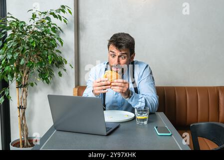 Giovane uomo d'affari che lavora a distanza dalla caffetteria durante la pausa pranzo. Lavoratore d'ufficio affamato che usa un computer portatile mentre mangia hamburger al restau Foto Stock