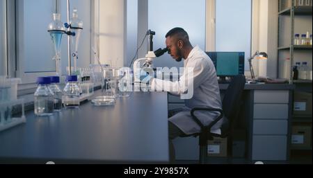 Laboratorio di scienza medica: Microbiologo maschio afroamericano guarda sotto microscopio, analizza campione di prova. Brillante scienziato utilizza apparecchiature high-tech. Una lavoratrice di laboratorio preleva le provette dal tavolo. Foto Stock