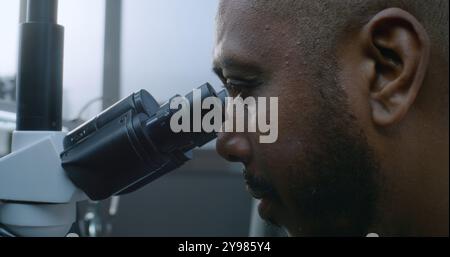 Flusso di lavoro in laboratorio di ricerca medica avanzata: Scienziato afroamericano maschile che utilizza il microscopio per sviluppare nuovi farmaci o formula di vaccino. Attrezzature di alta qualità e tecnologie moderne. Primo piano. Foto Stock