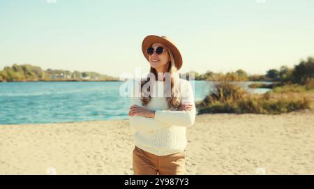 Donna matura ed elegante sulla spiaggia costiera, modella alla moda di mezza età che posa all'aperto, indossa maglione, cappello e occhiali, in piedi con il braccio incrociato Foto Stock