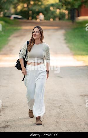 Una giovane donna passeggia tranquillamente attraverso un parco soleggiato, godendosi la libertà e la tranquillità della natura. Sembra rilassata e felice, circondata da vegetazione lussureggiante e alberi torreggianti Foto Stock