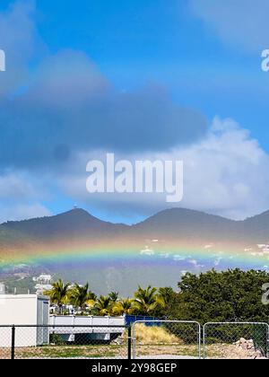 Un vibrante arcobaleno si innalza su lussureggianti montagne verdi, con una strada tortuosa che conduce attraverso il fogliame. Il cielo è azzurro e la scena è pacifica Foto Stock