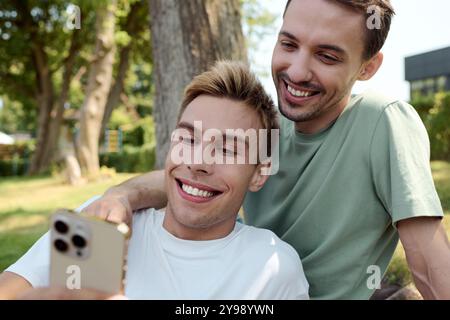 Una allegra coppia gay sorride al telefono, condividendo un momento delizioso nel parco. Foto Stock