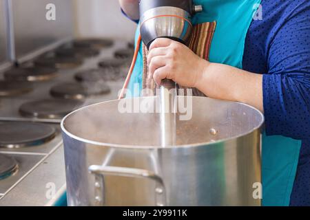 Cook utilizza un frullatore a immersione mentre prepara una grande quantità di cibo in una cucina industriale. Messa a fuoco selettiva Foto Stock