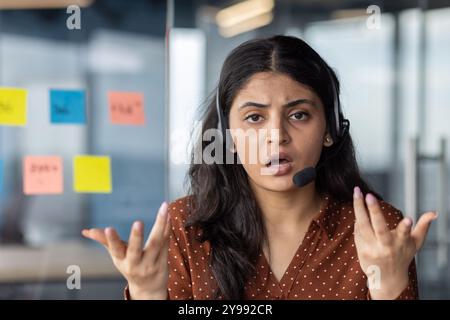 Un addetto al servizio clienti frustrato che indossa i gesti dell'auricolare con le mani, esprimendo confusione. Ufficio con appunti adesivi in background, che mostrano momenti di comunicazione difficili. Foto Stock