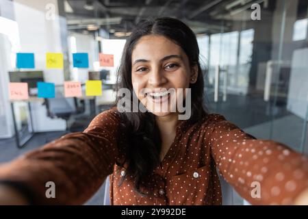 Una donna sorridente scatta selfie in un ambiente d'ufficio contemporaneo. Note adesive luminose sulle pareti in vetro esaltano la collaborazione. L'abbigliamento color legno contrasta l'elegante spazio di lavoro. Atmosfera ottimistica e coinvolgente Foto Stock