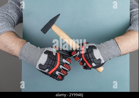 Braccio di un uomo nei guanti protettivi di un operaio edile o di un imprenditore edile che tiene il martello su sfondo di carta blu Foto Stock