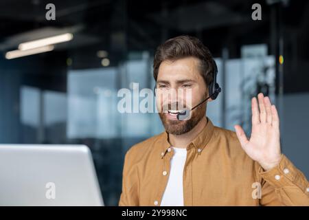 Uomo sicuro di sé durante una videochiamata in un ufficio moderno, con indosso la cuffia, impegnato nella comunicazione, salutando. Rappresenta il lavoro remoto, l'interazione professionale, le riunioni online, la connessione digitale. Foto Stock