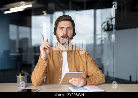 Uomo d'affari con cuffie e auricolari che prende appunti durante una riunione virtuale, sottolineando la comunicazione e la leadership nell'ambiente dell'ufficio. Persona che fa un punto, evidenziando una presentazione professionale. Foto Stock