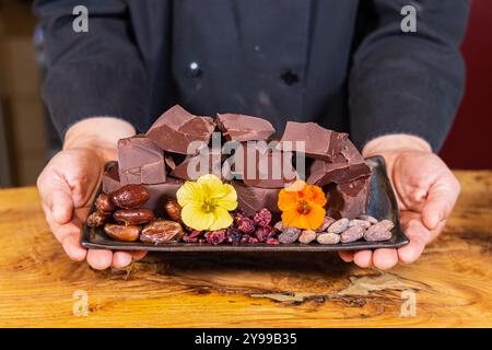 Cioccolataio con un vassoio di pezzi di cioccolato pregiati con fagioli di cacao, frutta secca e fiori commestibili. Messa a fuoco selettiva Foto Stock