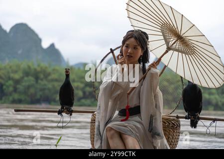 La ragazza di Hanfu seduta su una zattera di bambù tiene un ombrello mentre si fissa i capelli Foto Stock