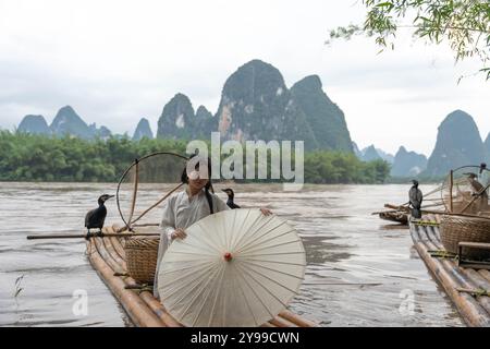 Una donna con abiti di Hanfu si alza il pollice tenendo in mano un ombrello su una zattera di bambù presso il fiume li a Xingping, Cina Foto Stock