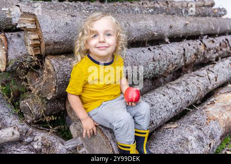 Il ragazzo con i capelli biondi ricci si siede sui tronchi, tenendo in mano una mela rossa, vestita come un piccolo boscaiolo in natura. Sorride felicemente, incarnando la gioia dell'infanzia. Messa a fuoco selettiva Foto Stock