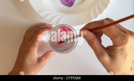 Il pennello ruota l'acquerello rosa in un bicchiere d'acqua trasparente, vista ravvicinata Foto Stock