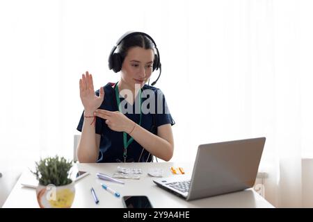 Allegro bella giovane medico donna in cuffie che posa al posto di lavoro in ospedale con computer portatile, guardando la fotocamera e mostrando caldo per controllare un p Foto Stock