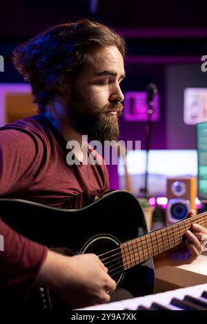 Musicista che crea una canzone suonando la chitarra acustica in studio domestico, registrando brani e cantando al microfono. Ingegnere del suono che mixa e padroneggia la sua musica con apparecchiature stereo e colonne sonore. Foto Stock