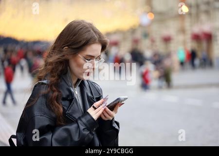 Ragazza attraente con occhiali da vista con il naso che pierca utilizzando lo smartphone per le strade della città, comunicazione mobile in autunno Foto Stock