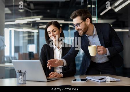 Colleghi in tuta che collaborano alla scrivania, concentrandosi sullo schermo del notebook con idee condivise. Uomo che tiene il caffè, donna sorridente, che indica il lavoro di squadra e il successo. Luminoso e moderno ambiente da ufficio. Foto Stock