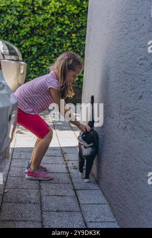 Amici divertenti: Bambina, bambina di cinque anni seduta a terra e gatto nero, per strada in Summertime. Foto di alta qualità Foto Stock