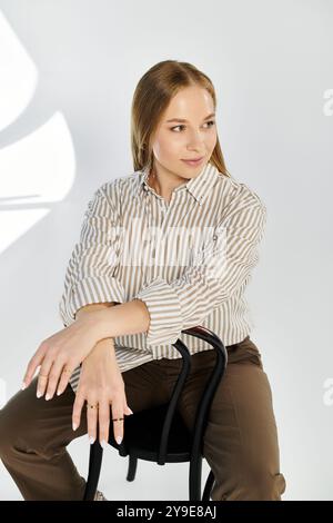 Una donna adorabile con capelli fluenti siede su una sedia, indossa una camicia e pantaloni a righe, trasuda eleganza in uno studio. Foto Stock