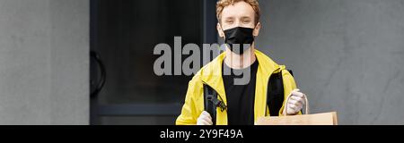 Un uomo di consegna che indossa una maschera medica nera porta un ordine in un edificio di uffici, garantendo la consegna sicura. Foto Stock