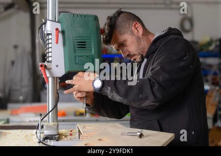 Uomo che ripara macchinari nella sua officina, con un trapano da tavolo automatico e abiti da lavoro neri Foto Stock