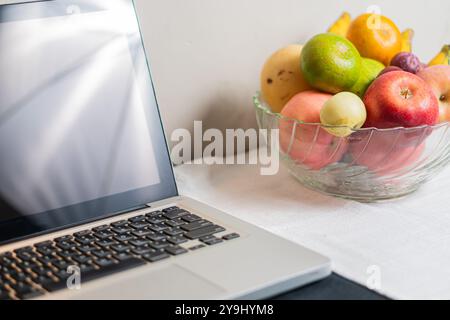 Un computer portatile moderno è seduto su un tavolo accanto a una ciotola di vetro piena di frutta, tra cui mele, pere e uva. Lo schermo del notebook è spento Foto Stock