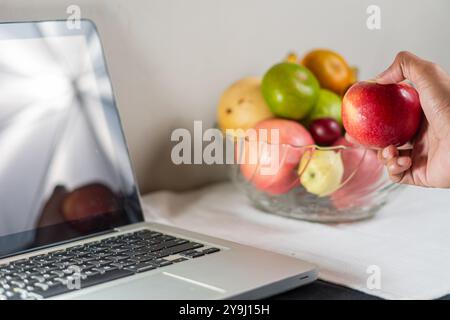 Una mano che tiene una mela rossa vicino a un computer portatile su un tavolo. Sullo sfondo, c'è una ciotola piena di vari frutti tra cui mele, pere e uva Foto Stock