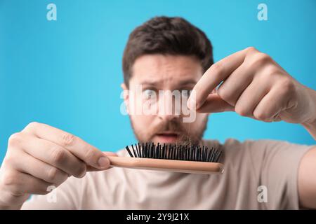 Uomo emotivo che prende i capelli persi dalla spazzola su sfondo azzurro, messa a fuoco selettiva. Problema di alopecia Foto Stock