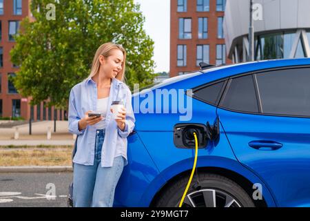 Giovane donna bionda che usa un'applicazione bancaria online per smartphone per pagare la ricarica di auto elettriche e bere caffè per strada. Foto Stock