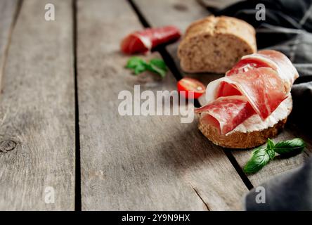 Concetto di cucina italiana. Pane tostato croccante con formaggio cremoso e prosciutto affettato sottile, pomodori ciliegini, rami di foglie di basilico, asciugamano da cucina su sfondo di legno. Pranzo in Italia, salutare pausa gustosa Foto Stock