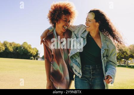 Due giovani fidanzate in un abbigliamento informale si godono una giornata di sole nel parco, ridendo e abbracciando, catturando l'essenza dell'amicizia e della felicità. Foto Stock