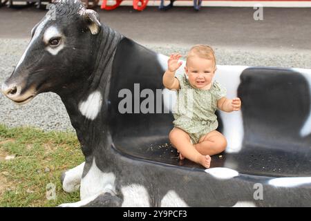 La bambina, in verde, è seduta sulla panchina a forma di mucca durante l'estate Foto Stock